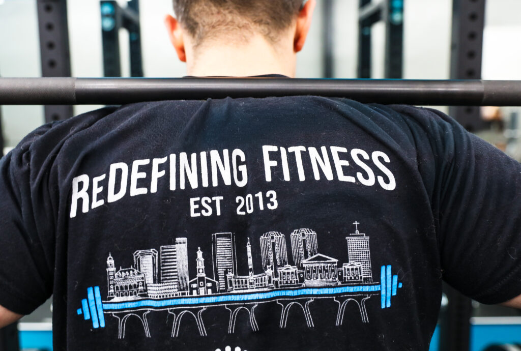 ReDefineRVA client under a barbell wearing a ReDefining Fitness Est 2013 shirt with the Richmond skyline